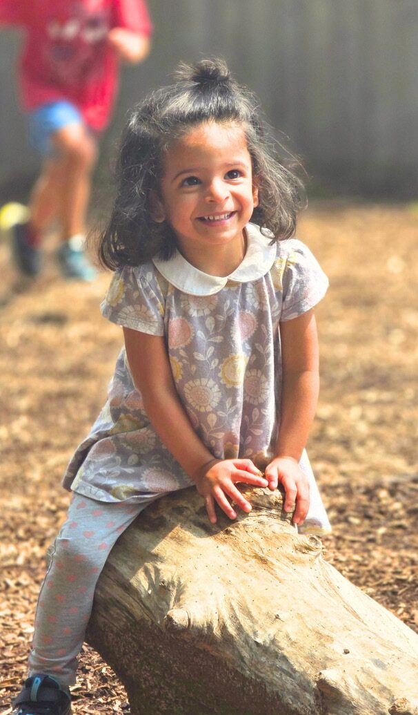 A Primary student sits on a log in the sun, smiling.