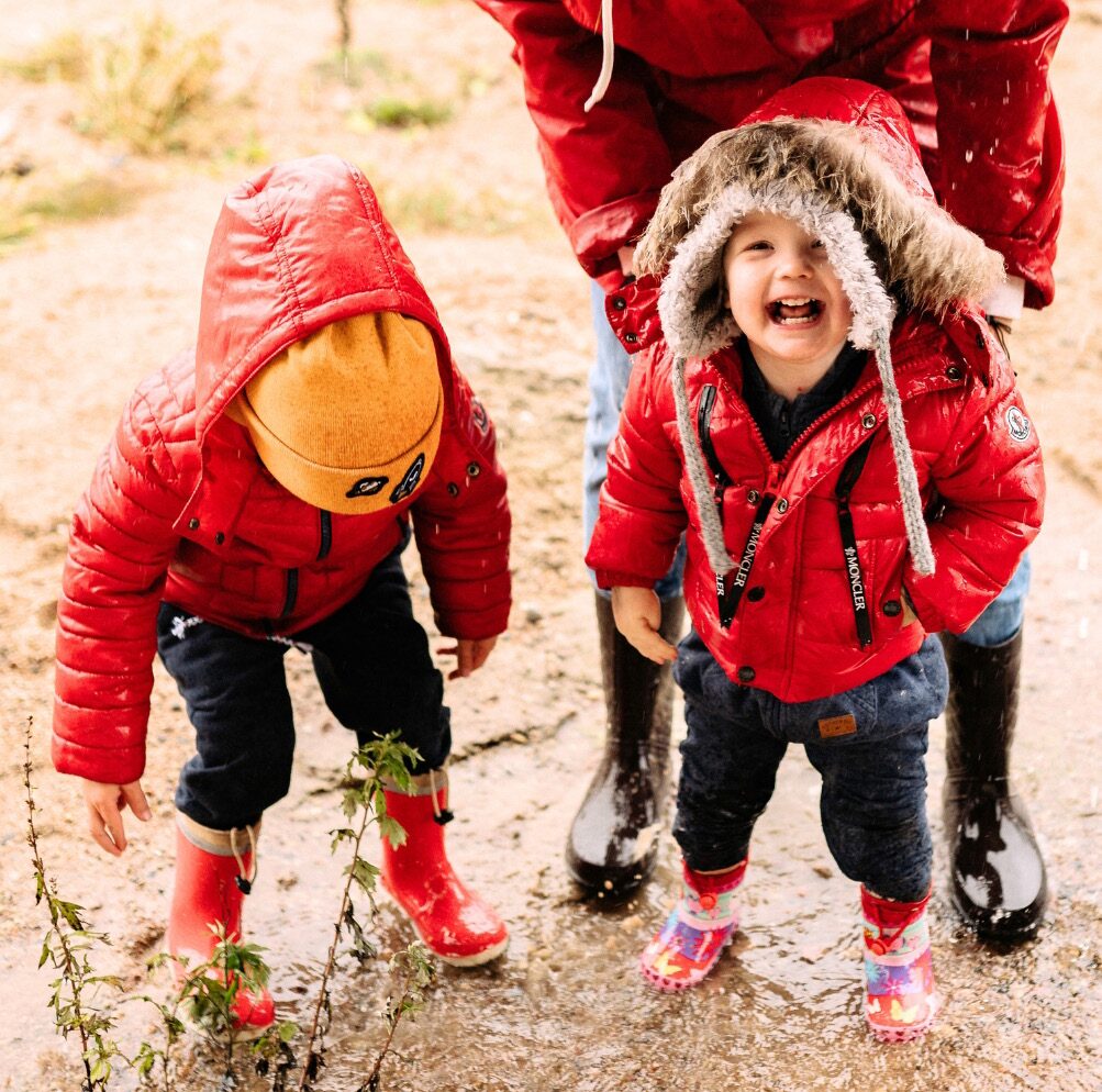 Two young children in red rain jackets and boots, laughing in the mud.